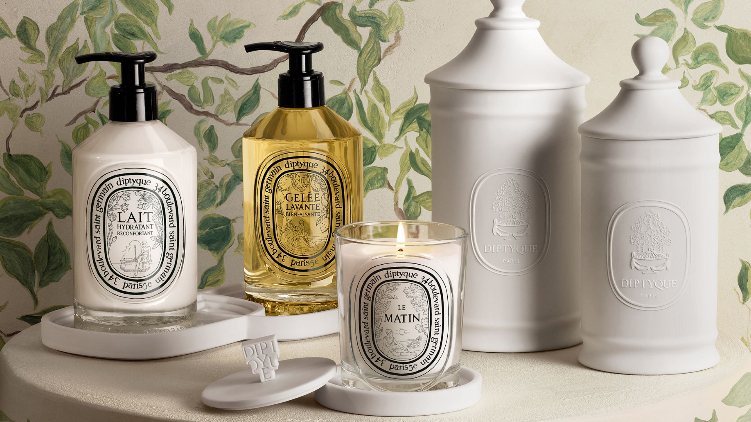 Diptyque Les Rituels de Soin collection on a white table with white wallpapered walls with vines