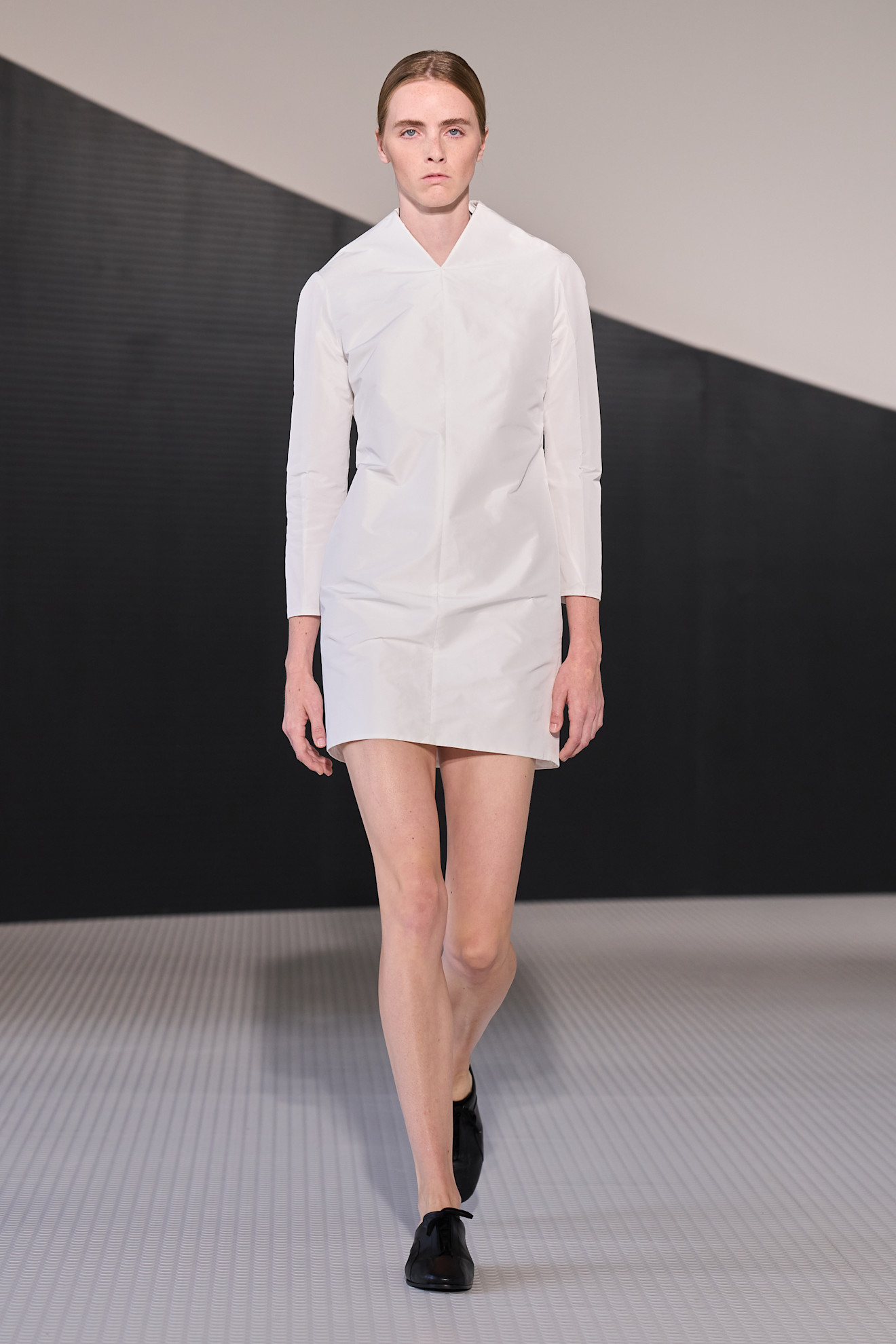 Jil Sander spring/summer 2026 model wearing a white jacket, mini skirt and black brogues.