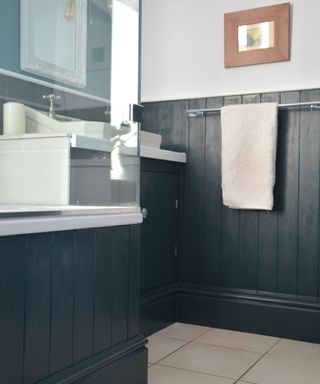 A bathroom with navy blue shiplap on half the walls, a glass shower, a white sink with a mirror, a silver towel rail with a white towel and a light gray wall with a rectangular wooden mirror above it