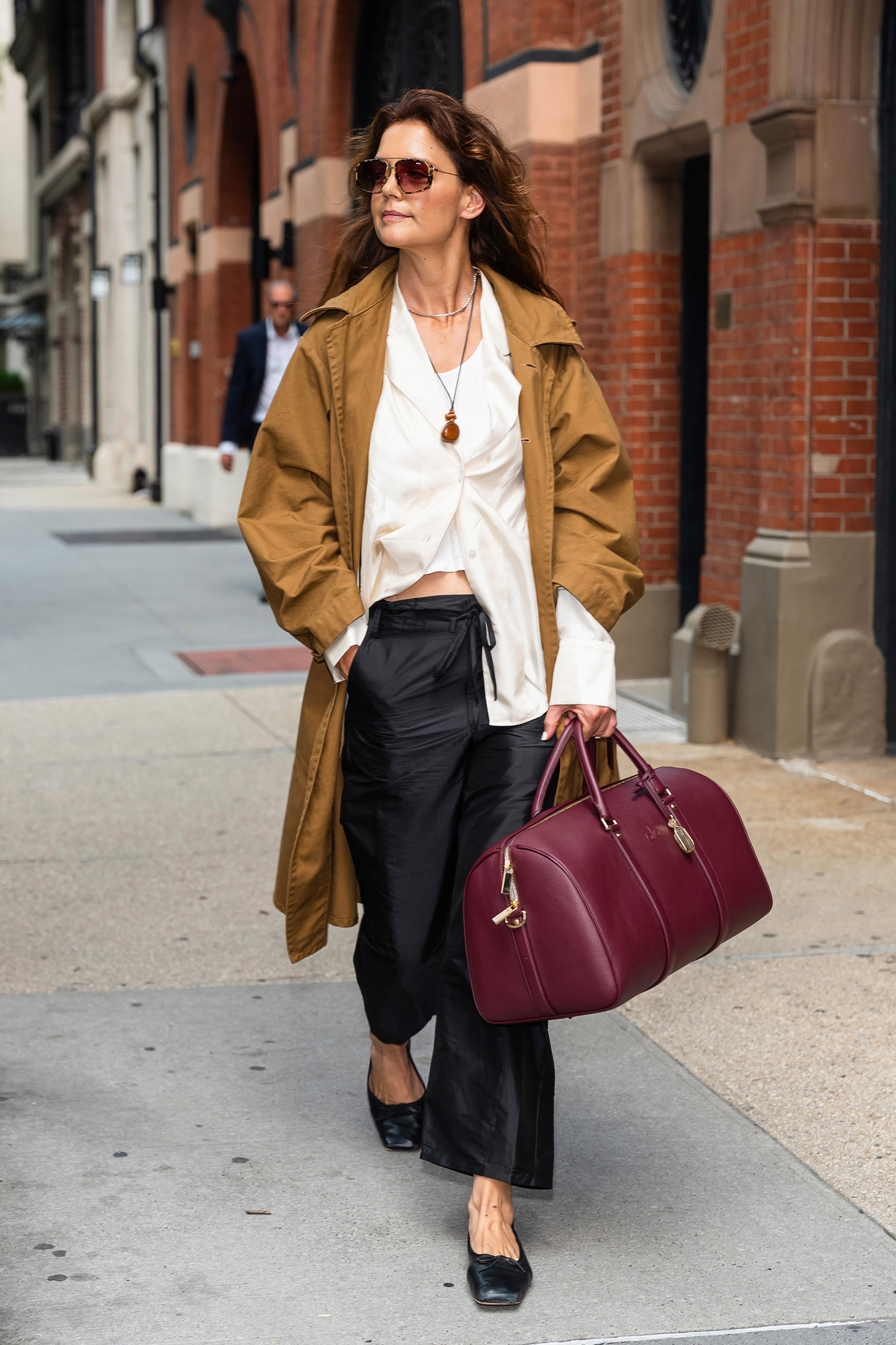 Katie Holmes wears a trench coat, white button-down, black trousers, black square-toe loafers, and a burgundy ALO bag.