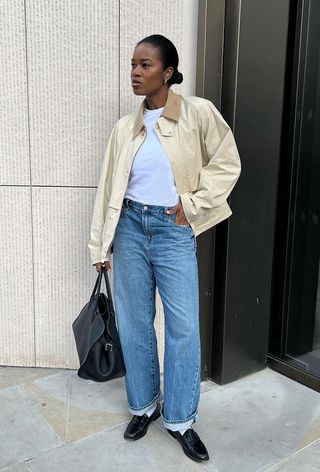 a photo showing the fall jacket trend, chore jackets worn by a woman standing against a white wall wearing a white barn jacket over a white t-shirt with jeans, black loafers, and a black bag