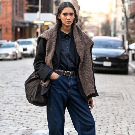 A woman wears a denim top, jeans, loewe runners, and a brown suede around her shoulders.