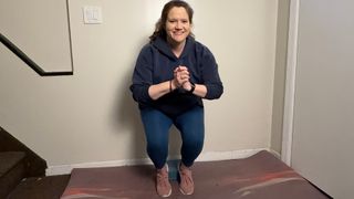 woman performing a squat against a wall with feet close together and heels up on a yoga block