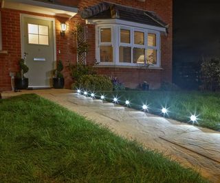 Warm white solar spotlights line a garden path leading up to a front door with an outdoor sconce light on a red brick wall