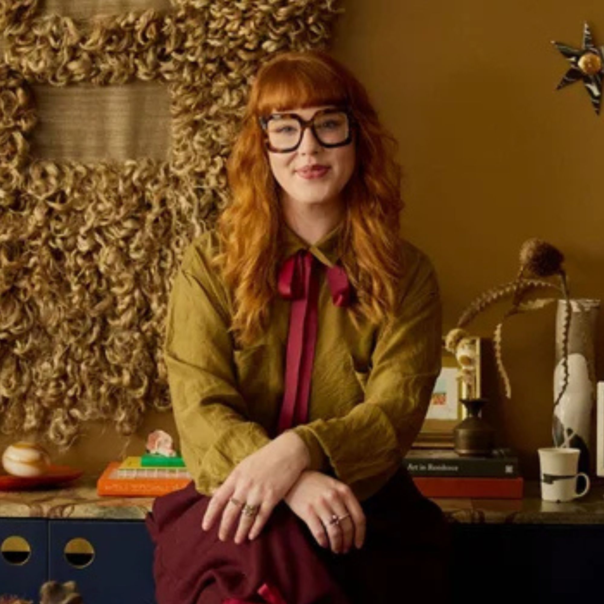 headshot of woman with red hair, fringe, and black-rimmed glasses in a colorful room with a rug on the wall