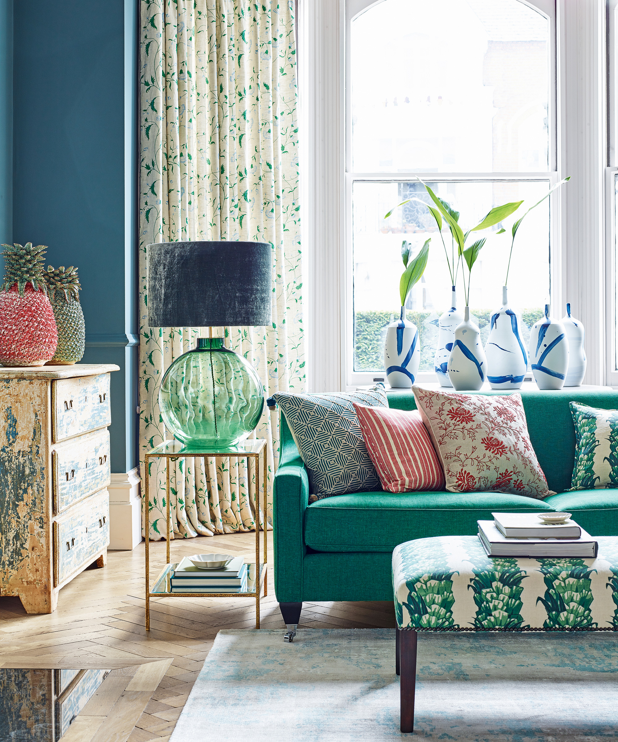 Living room trends illustrated by a green sofa with floral cushions, a large glass lamp and long floral drapes.