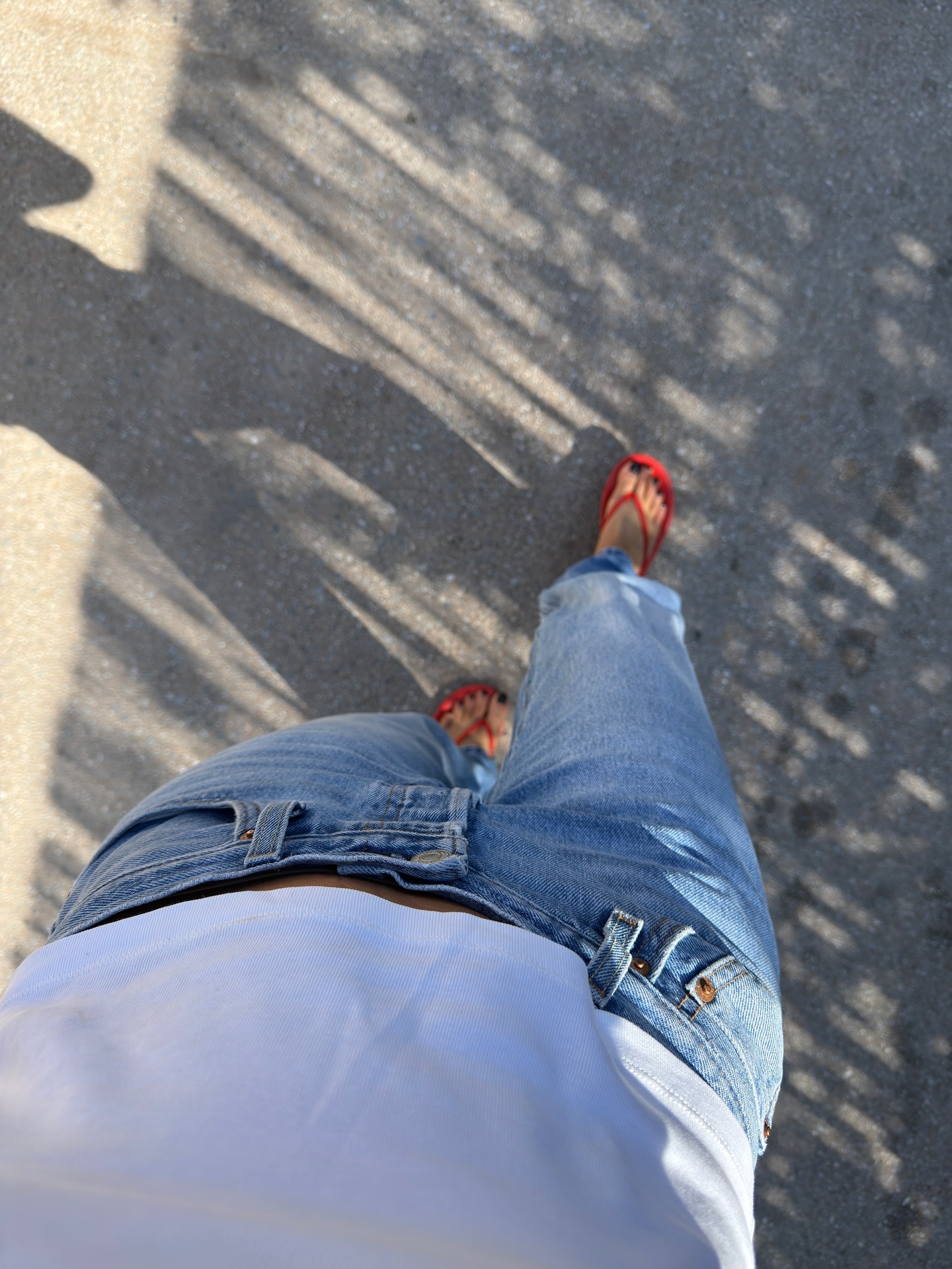 A woman taking a picture of her jeans and red flip flops with a black pedicure