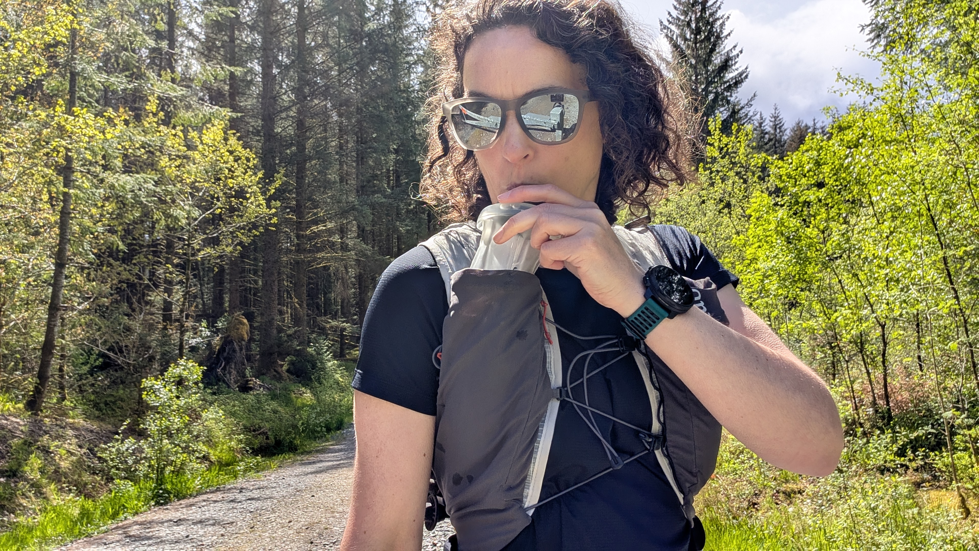 A woman trail runner wearing a black T-shirt, drinking water from a bottle in a grey Rab running vest