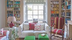 A vibrant living room with hardwood floor, a cream sofa with pink patterned cushions, two green pouffes, a striped armchair with brown wooden handles, and a small circular table with a green and white leafy tablecloth and burgundy lamp with red and white lampshade. Behind is a large square window with green and white blinds, flanked by floor to ceiling bookshelves filled with color-coded books.