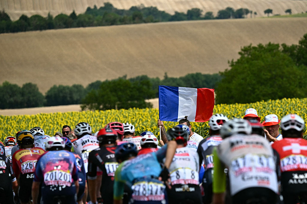 The pack of riders (peloton) cycles past a spectator waving a French flag during the 15th stage of the 112th edition of the Tour de France cycling race, 169.3 km between Muret and Carcassonne, southwestern France, on July 20, 2025. (Photo by Loic VENANCE / AFP) (Photo by LOIC VENANCE/AFP via Getty Images)