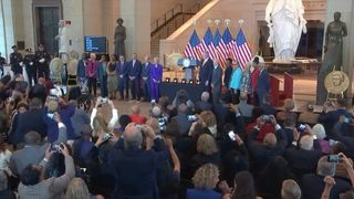 a dozen well-dressed people stand on a stage, with five american flags in the background