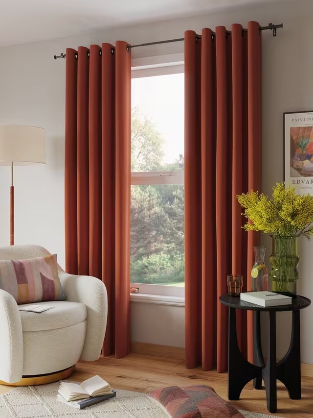 living room with burnt orange eyelet curtains over window, a beige arm chair with colorful cushion, a floor lamp behind the chair, a white rug with a book open on the floor, another cushion on the floor, a black side table styled with a book, glass, jug, and vase of flowers and an artwork on the wall behind