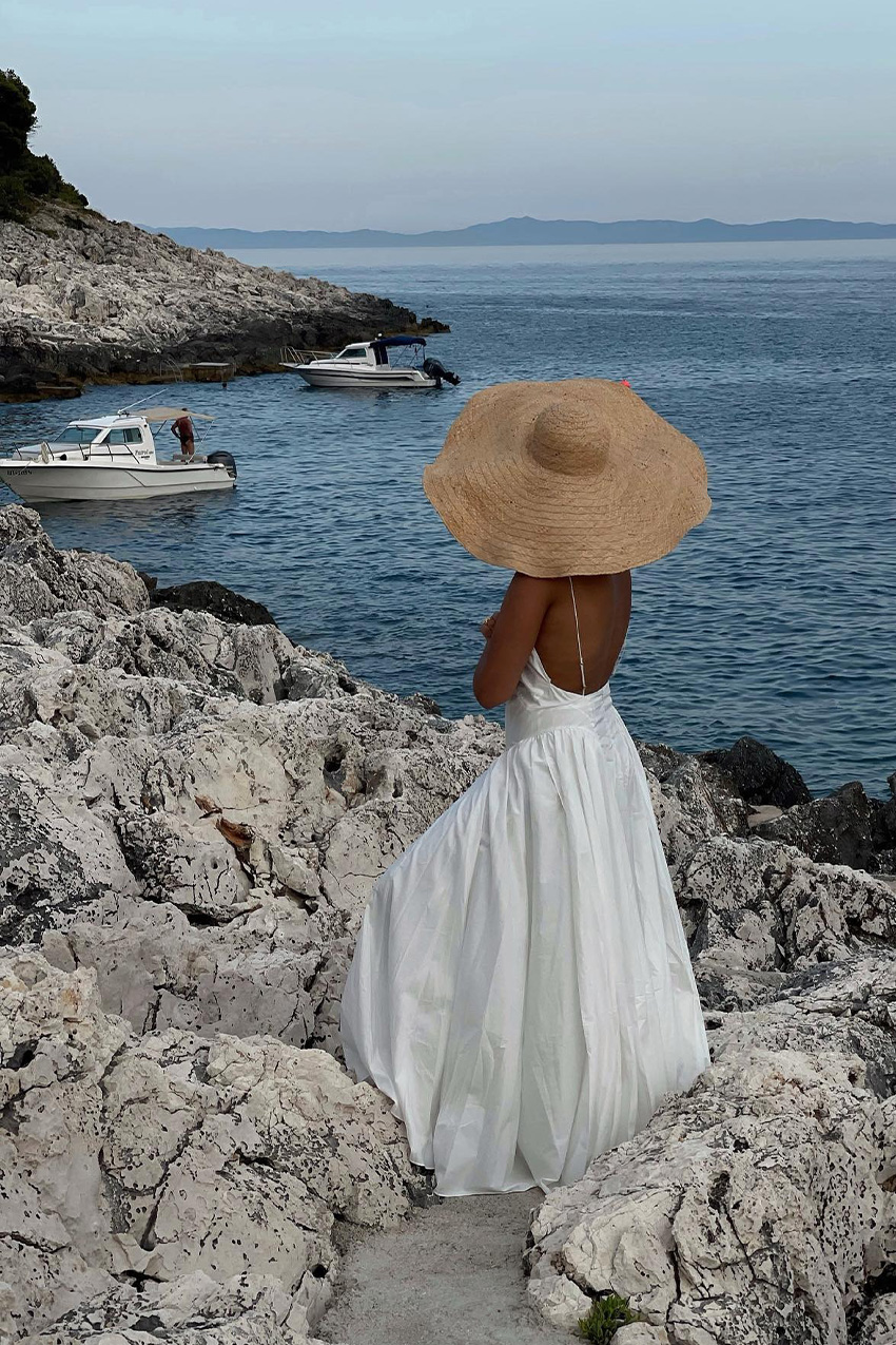 European vacation outfit idea is shown in a photo of a woman standing by the sea wearing a raffia woven hat, a white maxi dress, and flip flops