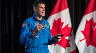 jeremy hansen on stage in an astronaut flight suit. he holds a passport with his left hand and gestures to the audience with his right. behind are two large canadian flags