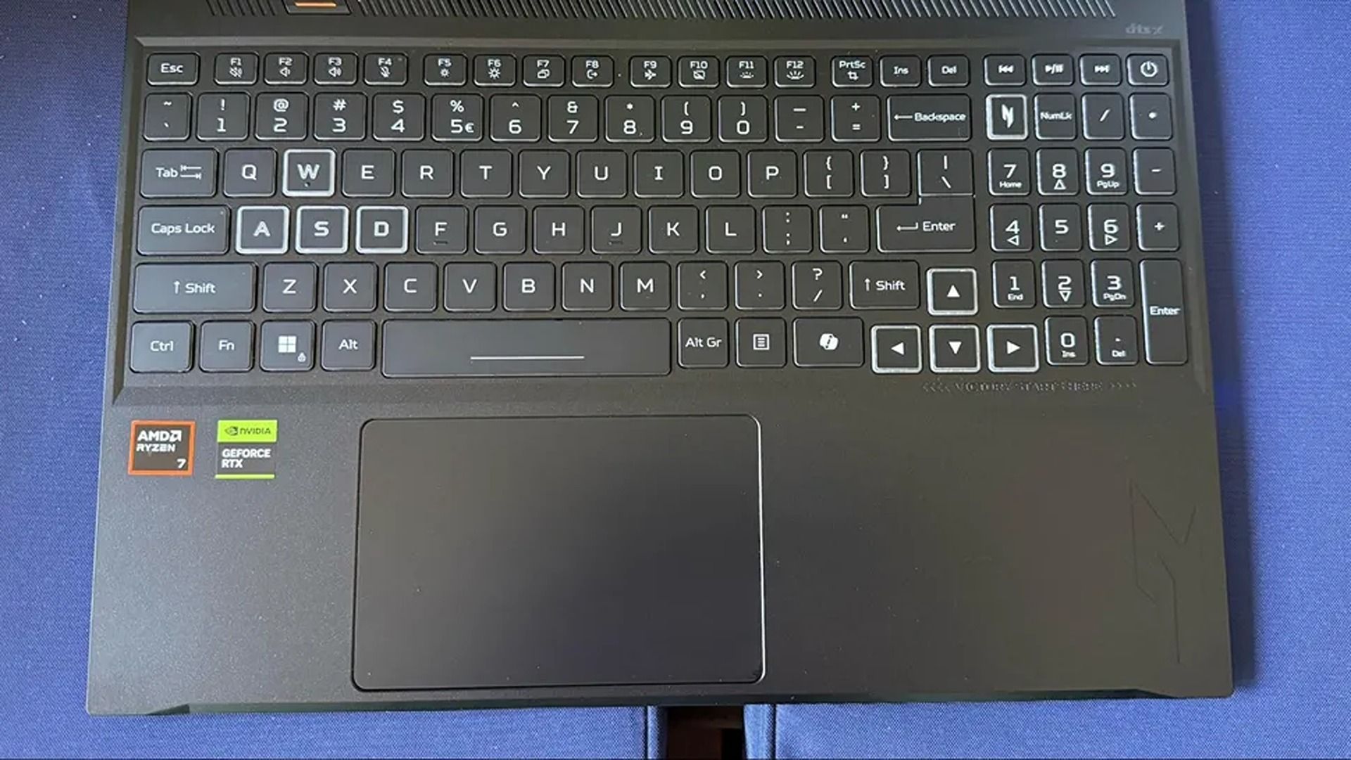 Top-down view of a black laptop keyboard and large touchpad on a blue surface, with AMD Ryzen and Radeon stickers at the lower left; practical, utilitarian design.