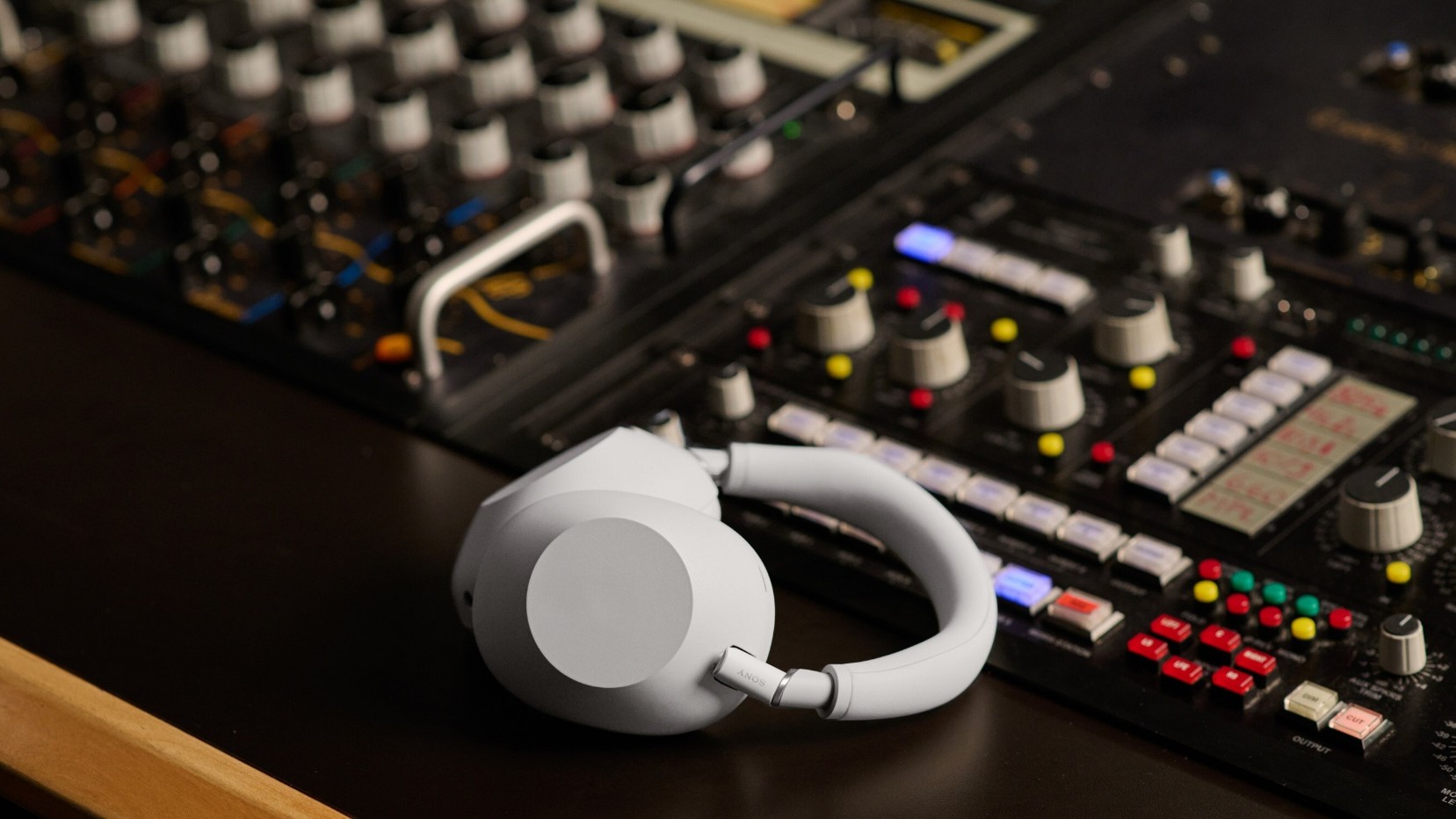 A pair of Sony's newest headphones in white sitting on in a music studio against a soundboard.