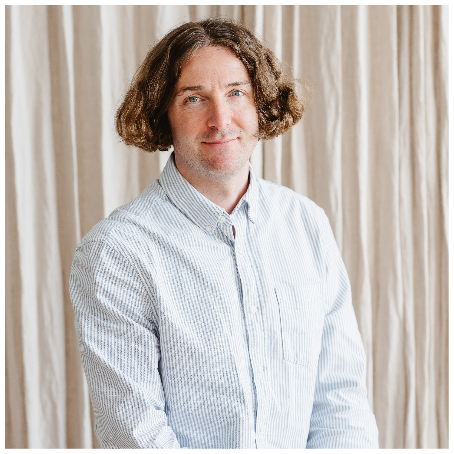 A headshot of a male with cheek-length brown hair in a blue striped white shirt against a curtain backdrop