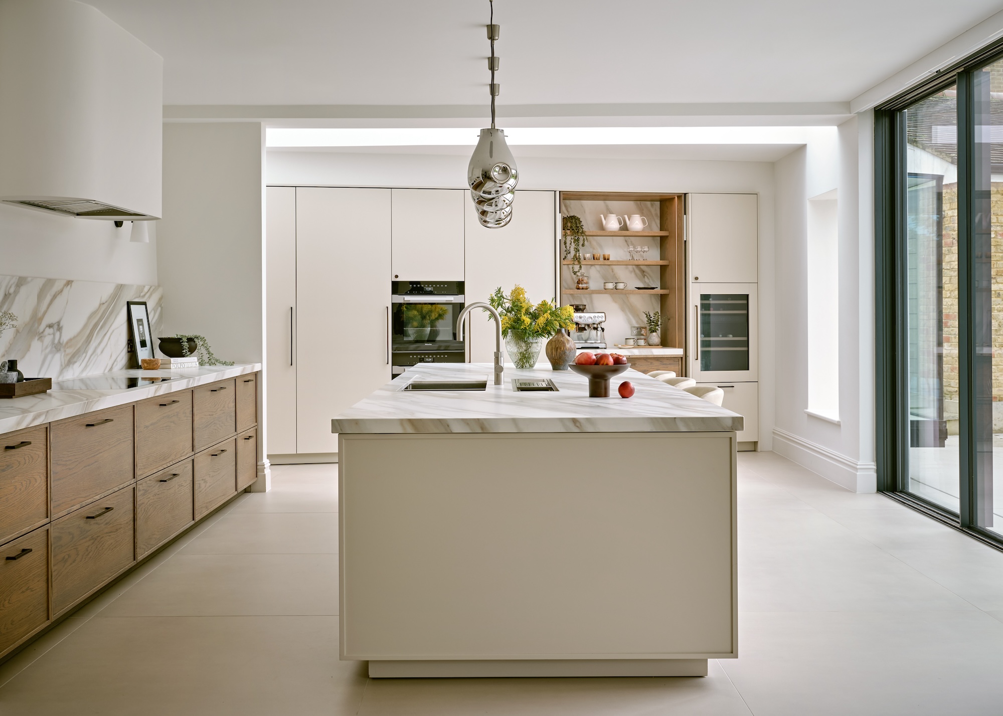 A zoned kitchen layout in off white and wood, consisting of a central island unit, a run of base units and a wall of floor-to-ceiling storage