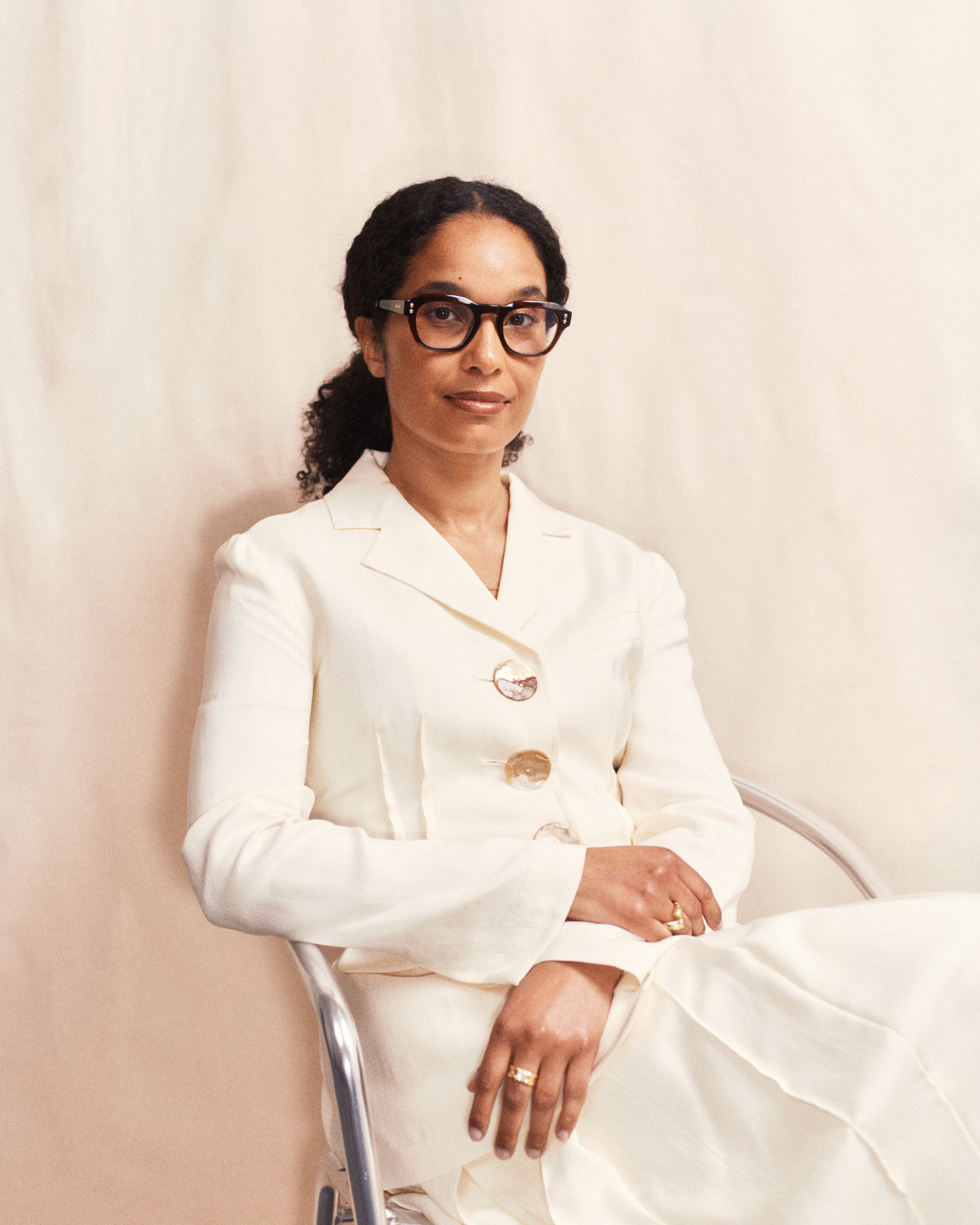 A young woman dressed in a white, loose-fitting suit with jewel mother of pearl buttons wears a pair of brown glasses and golden jewelry while sitting on a chrome chair in front of a beige background.