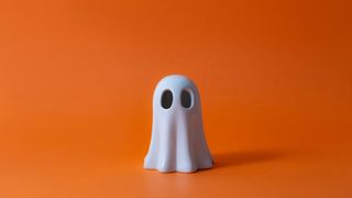 A small plastic cartoon-style ghost, sat on an orange background. The ghost resembles a white bedsheet with black eyeholes cut at the top.