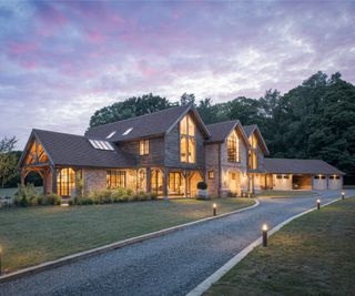 A large barn-style oak frame self build clad in timber and brick on a large rural plot with trees in the background, and with substantial garages to one side