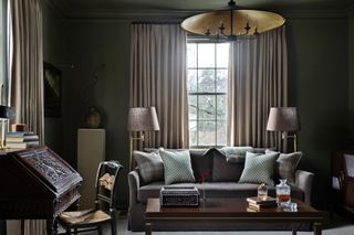 dark living room with gold pendant light, curtains over windows, a dark sofa with lots of throw pillows and lamps on either side, a writing desk with chair, a plinth holding a vase in the background, and a timber coffee table