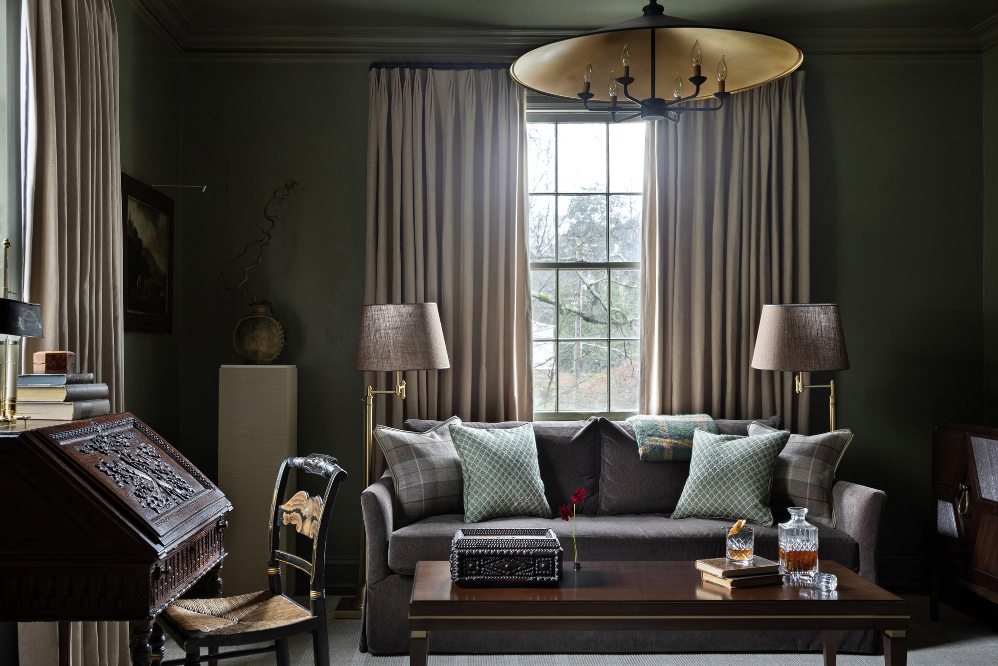 dark living room with gold pendant light, curtains over windows, a dark sofa with lots of throw pillows and lamps on either side, a writing desk with chair, a plinth holding a vase in the background, and a timber coffee table