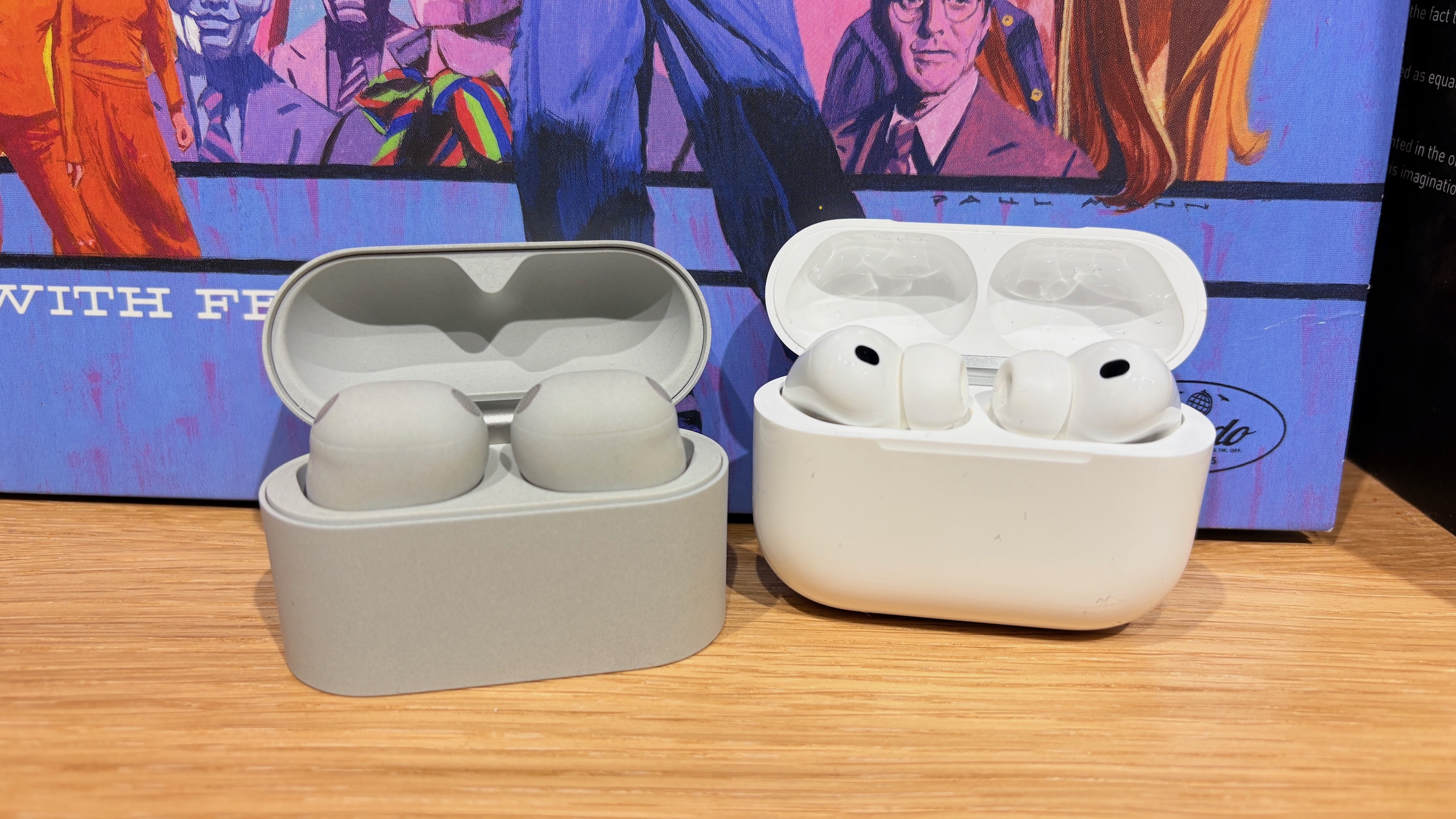 The Sony WF-1000XM6 and Apple AirPods Pro 3 wireless earbuds in their open charging cases on a wooden surface.