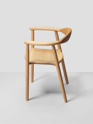 A wooden chair with thin legs leading up to two curved arms. The back is open with a small curved back support piece. The seat base is wooden with side inward curvatures and front/rear outward curvatures.