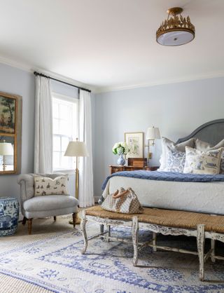 double bed with pale blue walls and blue bed linen and headboard oriental style rug and antique bench at foot of bed and pale blue nursing chair with cushion and pale curtains