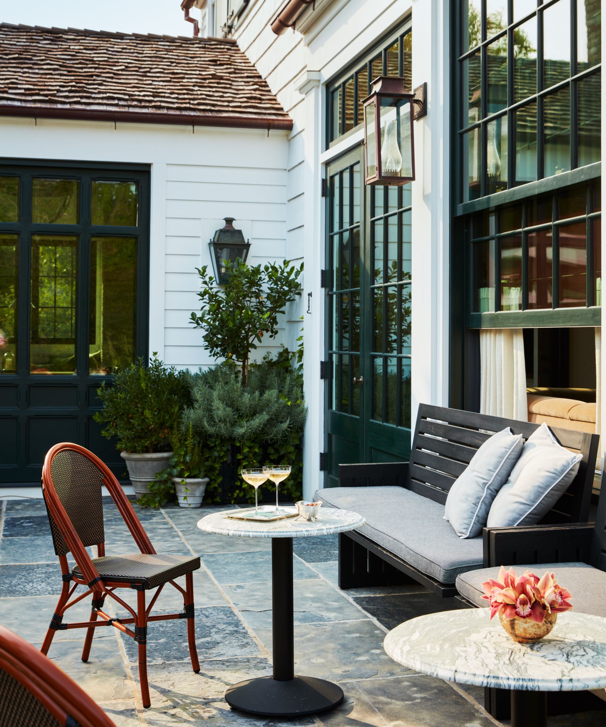 patio space next to a house, with some plants, black bench and grey cushions, a marble round table with cocktails on it and a chair