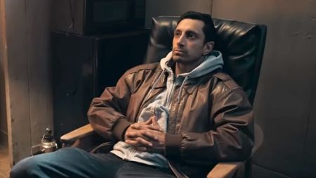 Riz Ahmed sitting back in a chair looking ahead in Relay. 