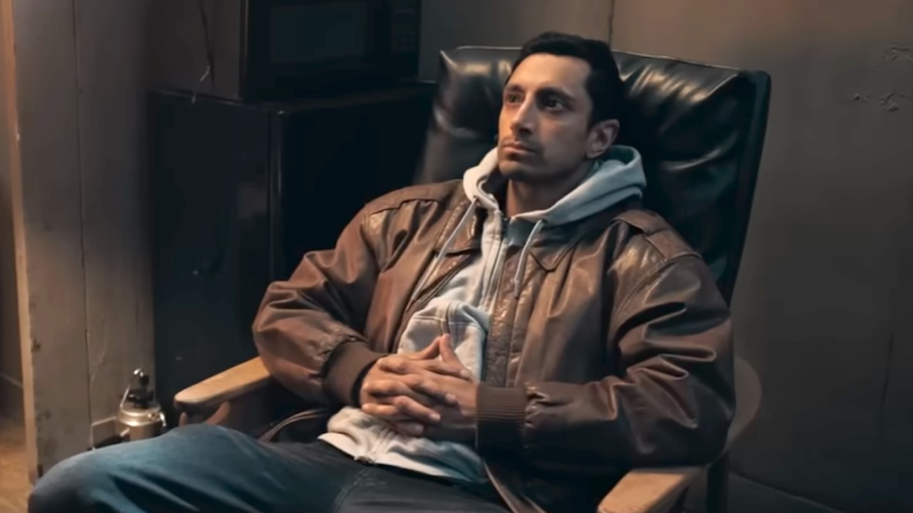 Riz Ahmed sitting back in a chair looking ahead in Relay. 