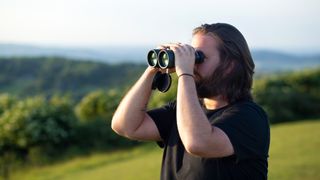 Jason Parnell-Brookes holding a pair of Celestron Nature 12x56 binoculars outside with rolling hills in the background