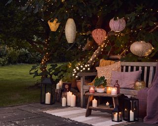 Relaxed boho garden scene with bench and colored cushions, candlelight staggered on ground, twinkling fairy lights around tree trunk, and intricate colored lanterns hanging from tree.