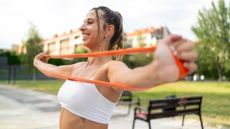 woman facing sideways to the camera pulling apart an long loop orange resistance band across her chest. she's in a park setting. 