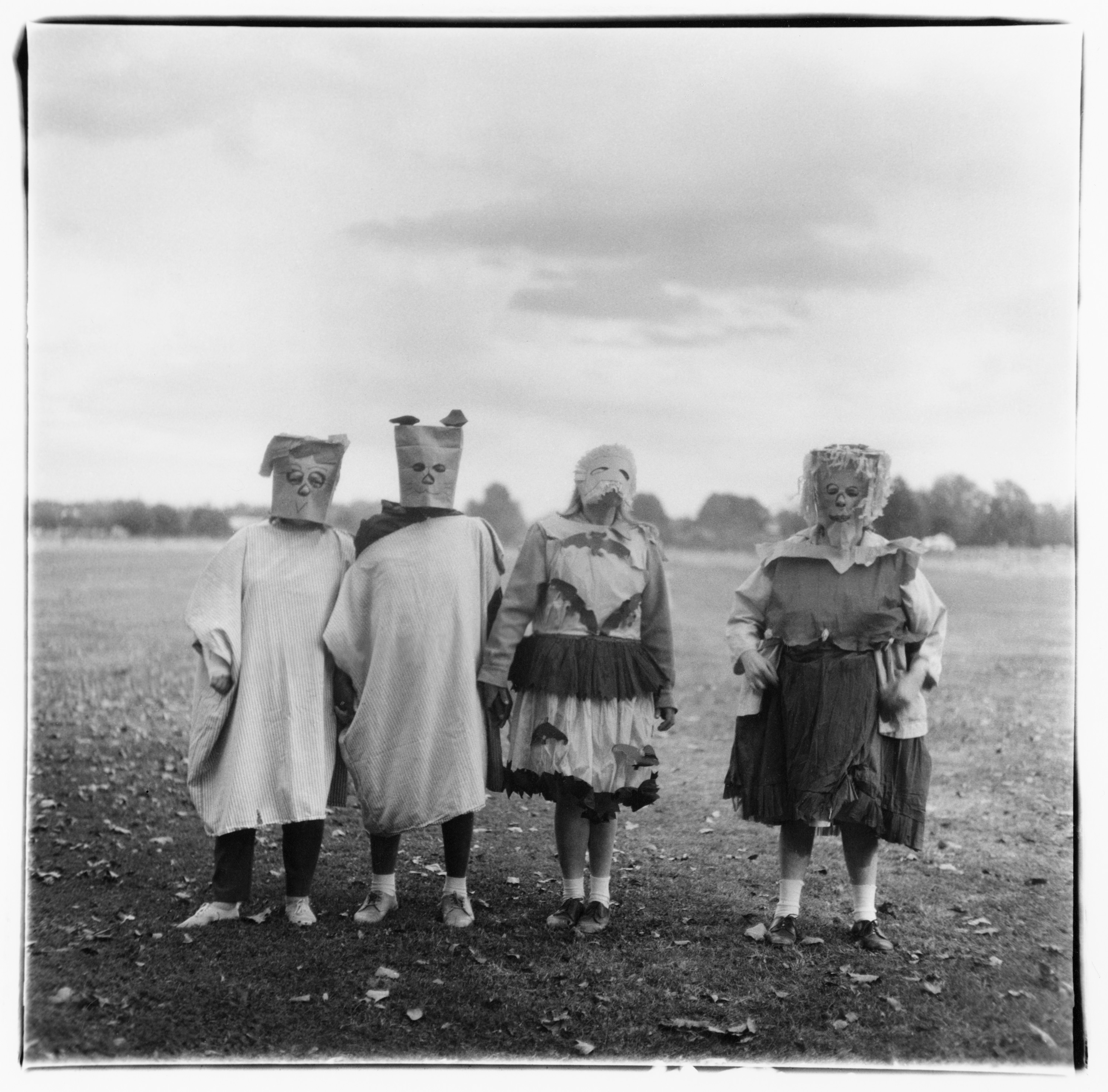 Four figures in unsettling Halloween costumes, two with crude paper-bag masks and two with ragged face coverings, stand side-by-side in a field.