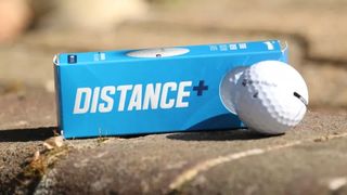 TaylorMade Distance+ Golf Ball and its blue packaging resting on a wall on the golf course