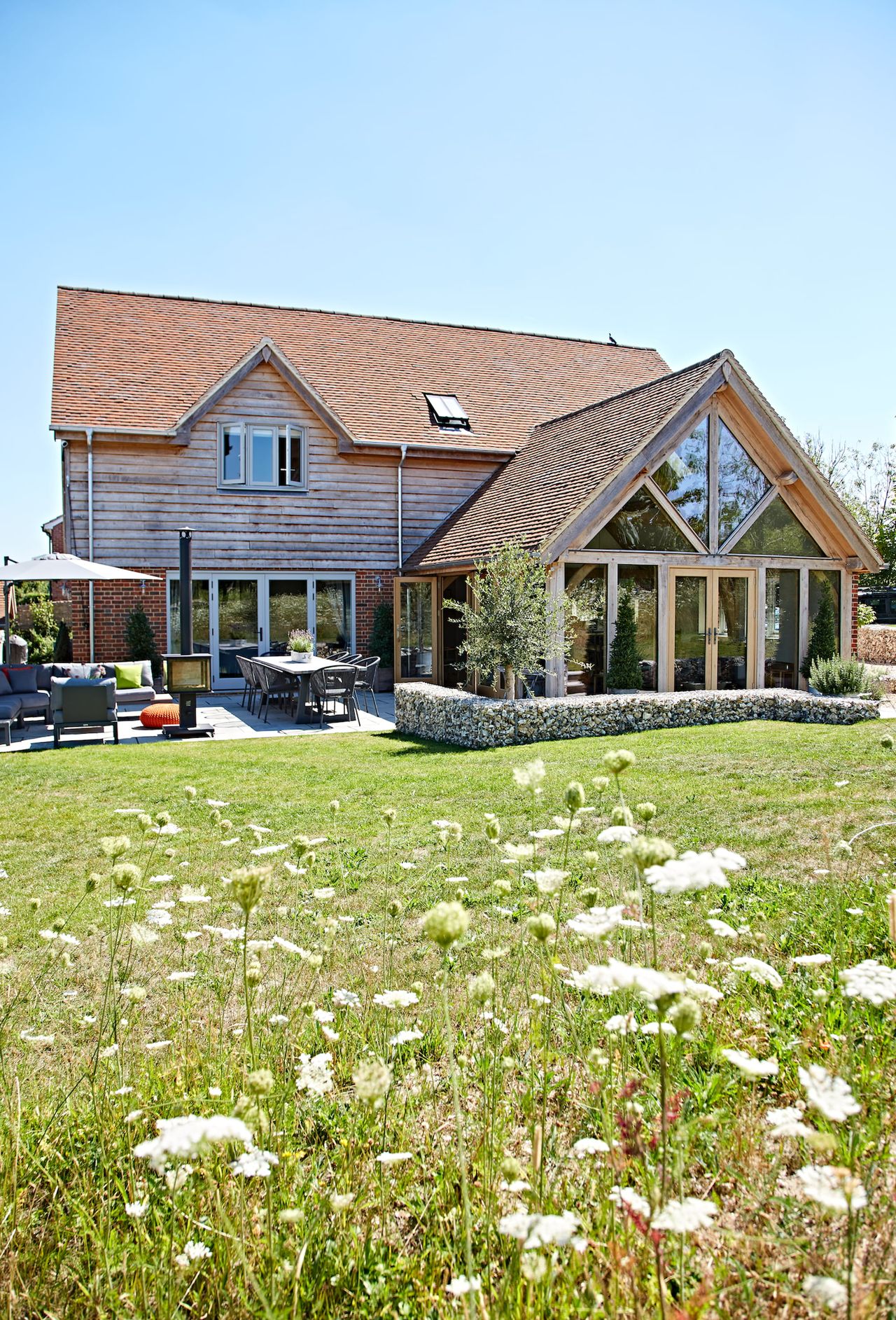 A modern rustic house with a glass and oak frame extension surrounded by meadow gardens