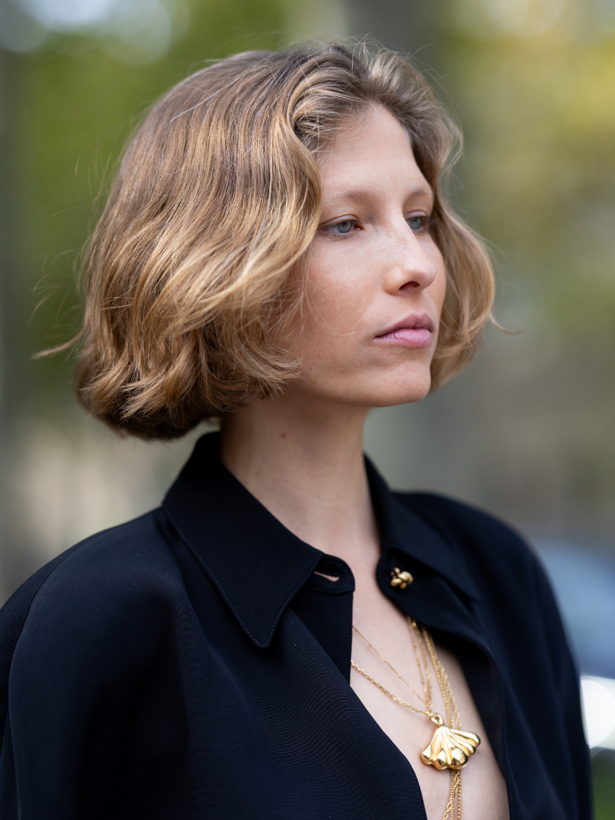 Katharina Kaminski wears layered gold necklaces with shell pendant, a black crepe plunging shirt with gold knot button