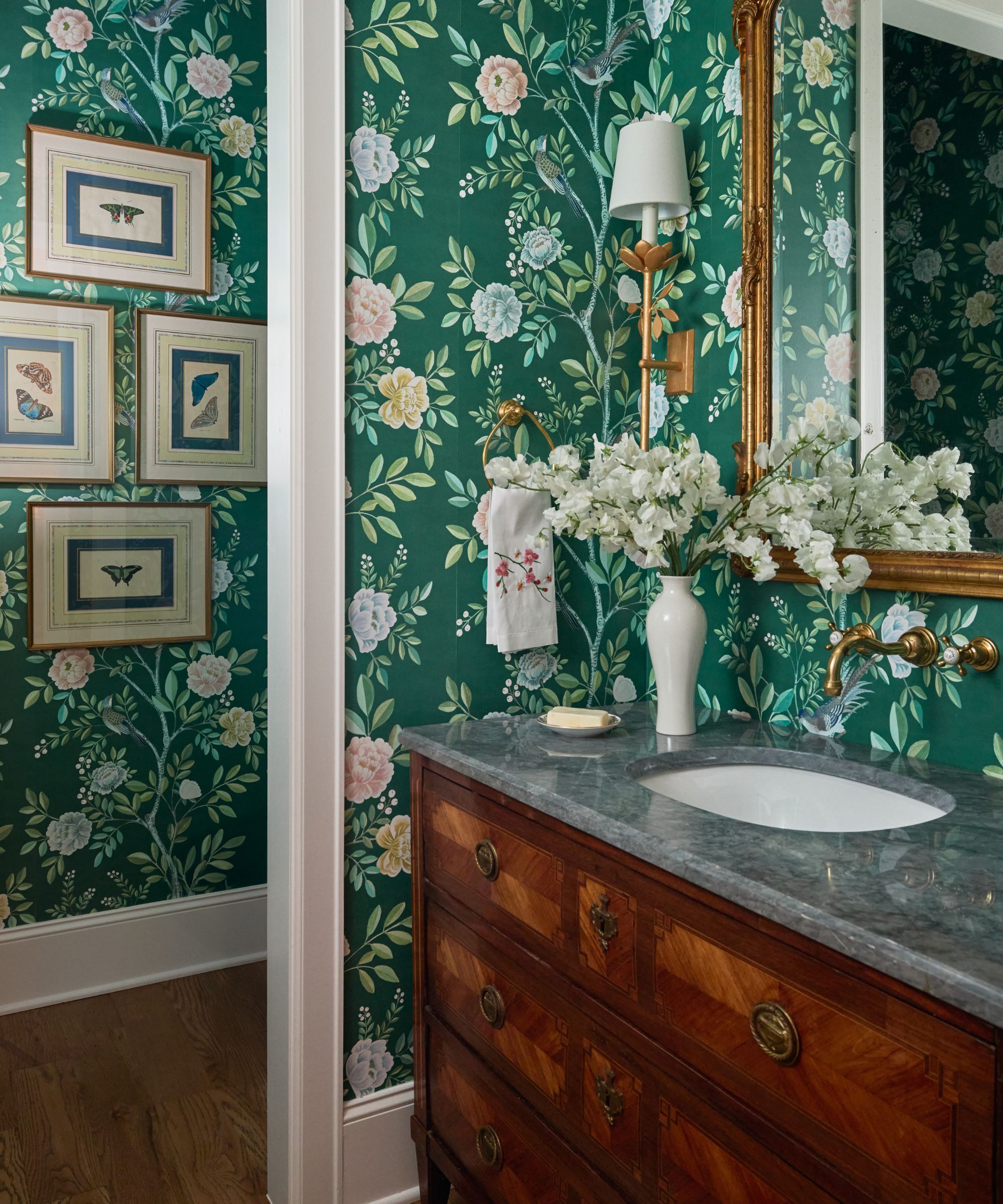 Elegant bathroom with green floral wallpaper, a wood vanity with a grey marble top, brass wall sconce, gold-framed mirror, and framed butterfly prints on the wall.
