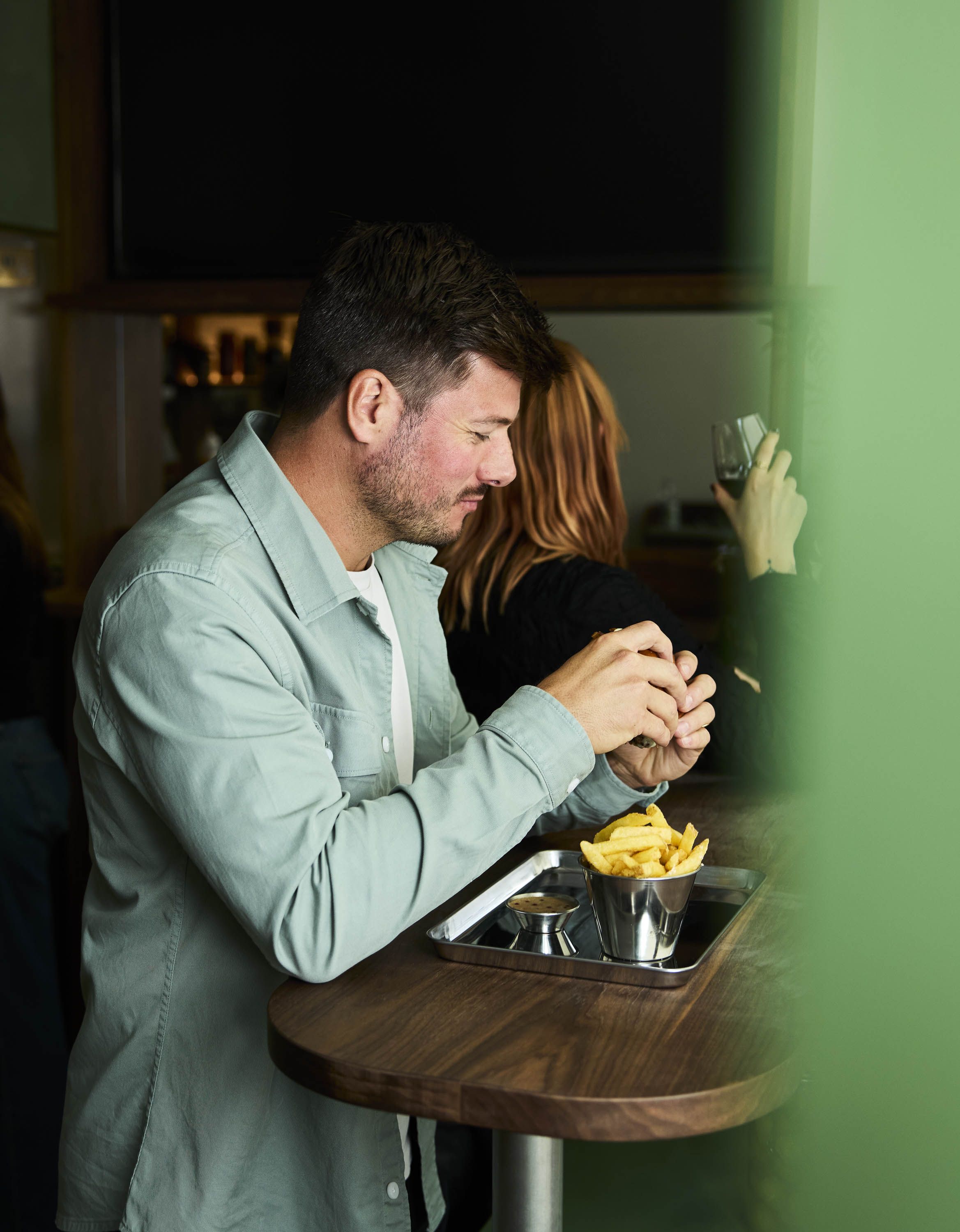 A burger joint's neon-lit sign, a man eating fries and a burger inside it, and a close-up of a chrome platter filled with a large burger and a portion of fries, paired with a glass of wine that's just been poured for the diner.