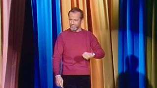 George Carlin in a mauve shirt performing in front of orange and blue curtain, footage featured in HBO's doc George Carlin's American Dream.