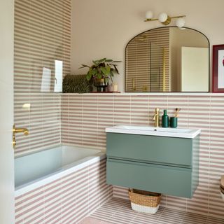 Bathroom with pink and white striped tiles on the wall and the bath, with a green vanity unit and a mirror above it