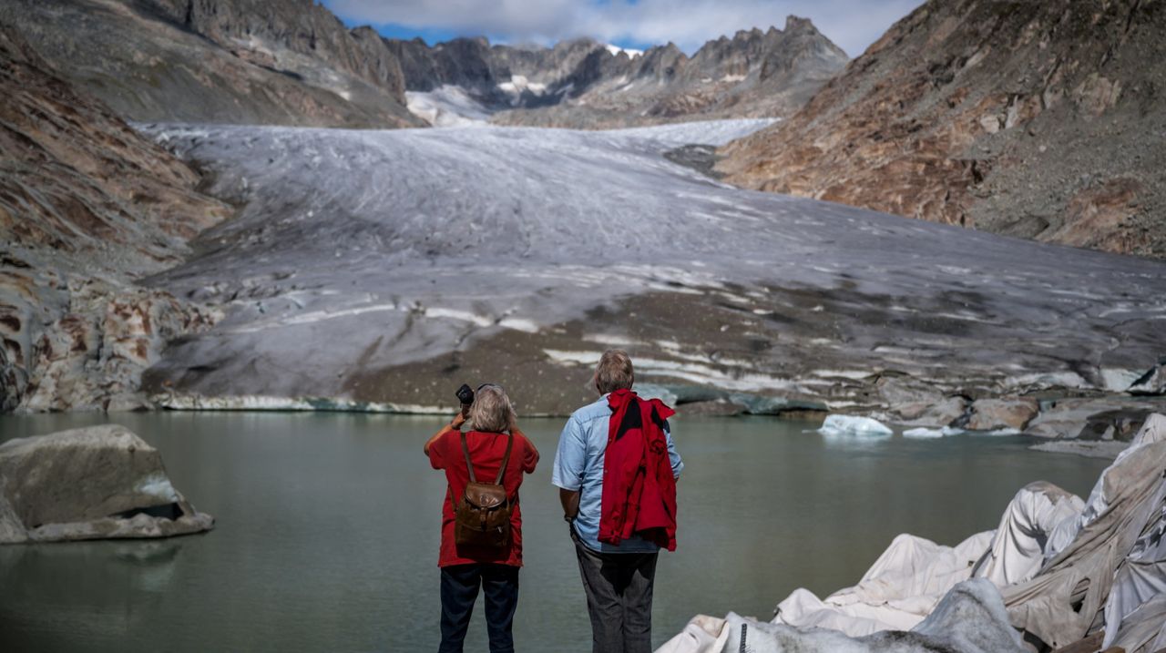 This photograph taken on September 12, 2025 above Gletsch, in the Swiss Alps, shows two tourists facing the Rhone Glacier melting into its glacial lake. Switzerland's glaciers, which are disproportionately impacted by climate change, have shed a quarter of their mass in the past decade alone, a study warned amid concerns the melt is accelerating. In 2025, glacial melting in Switzerland was once again "enormous", the Glacier Monitoring in Switzerland (GLAMOS) network said, adding it was close to the record set in 2022. (Photo by Fabrice COFFRINI / AFP) (Photo by FABRICE COFFRINI/AFP via Getty Images)