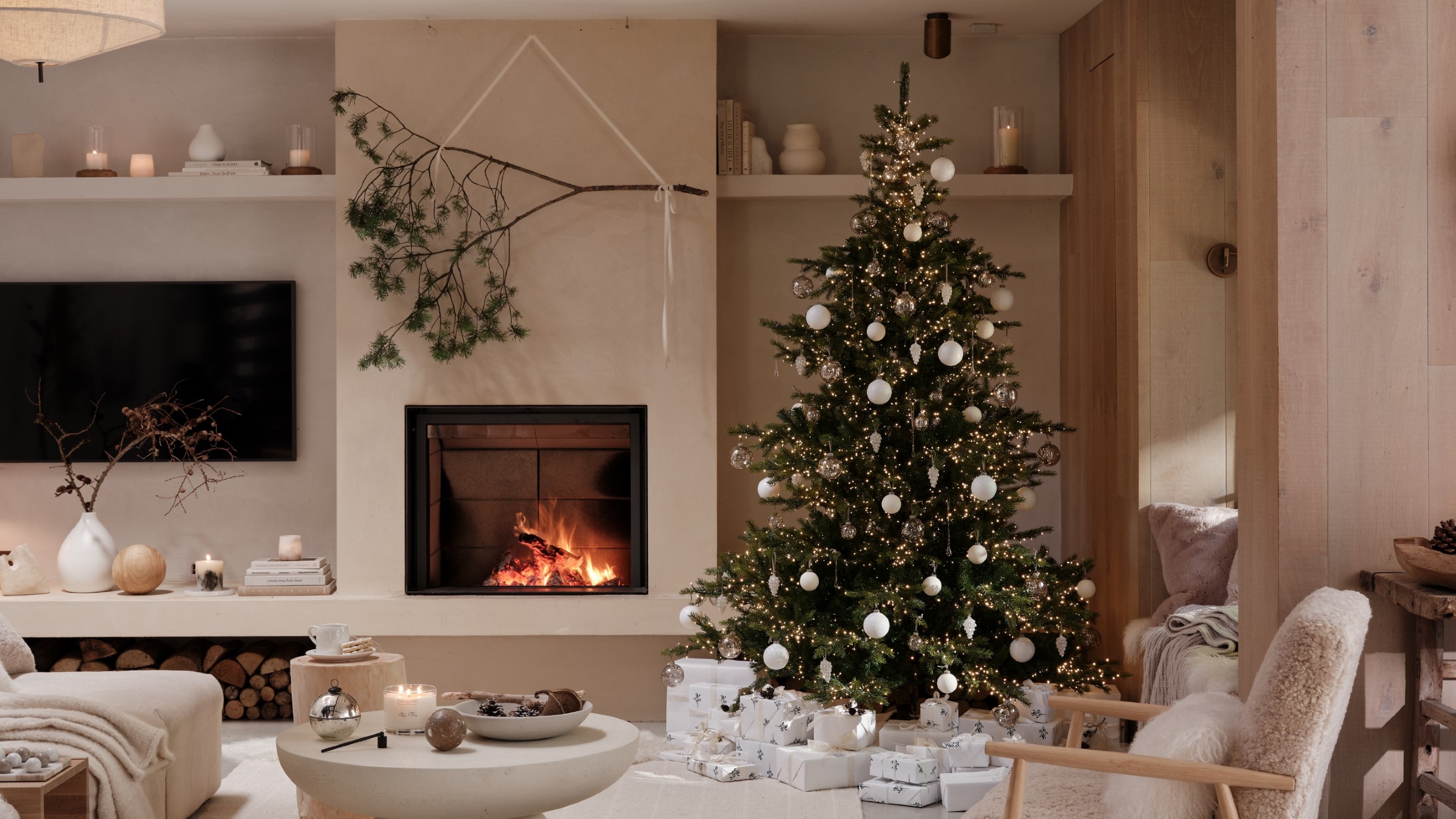 The White Company's extra bright Christmas tree in a modern living room with a lit fireplace and a decorative branch hung above it