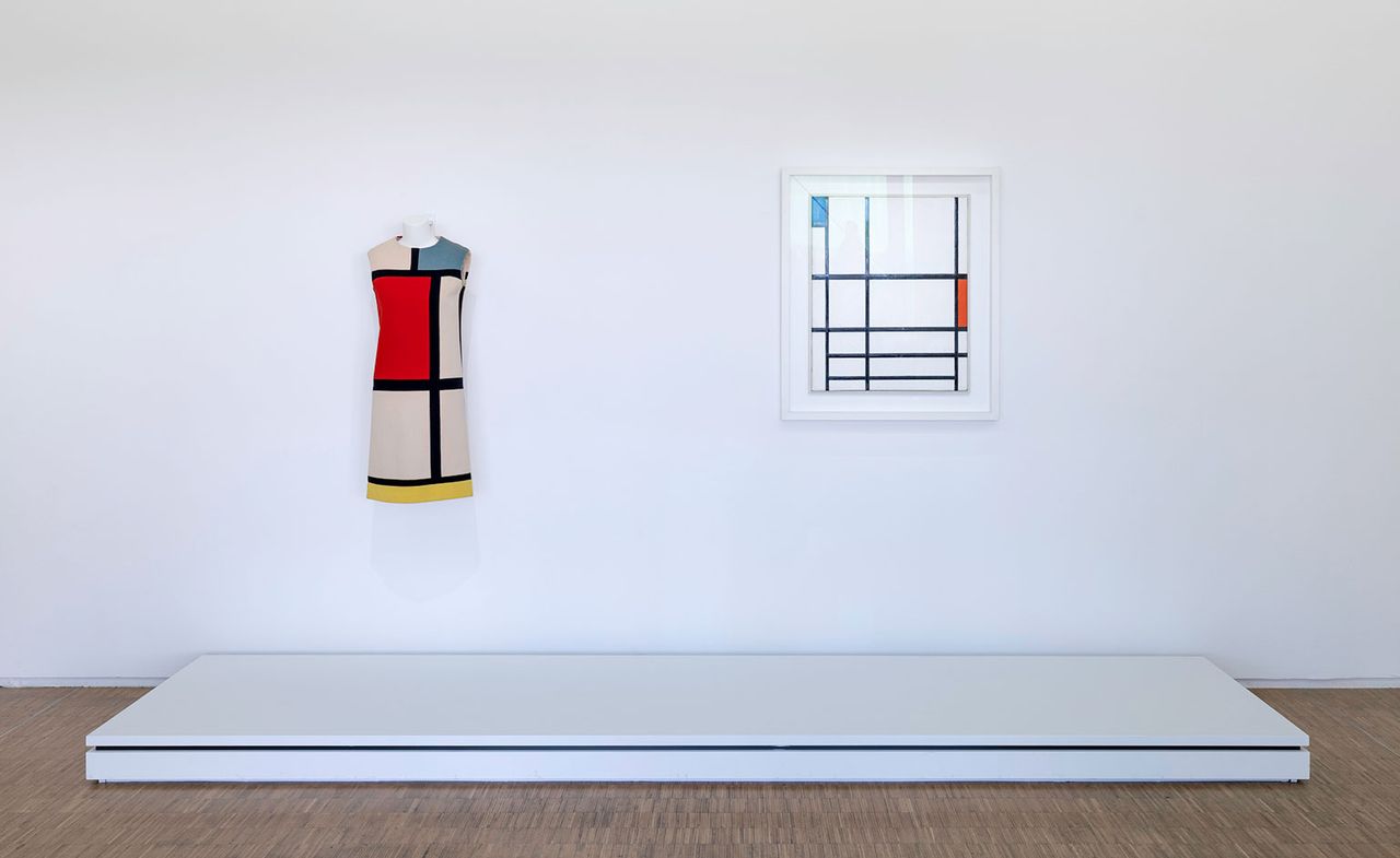 View of Centre Pompidou&#039;s tribute to Yves Saint Laurent featuring a colour block dress and wall art on display in a room with white walls, a white platform and wood flooring