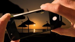 Two hands holding an iPhone 16 taking an image of a sunset over the ocean with parasols in the foreground