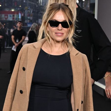Sienna Miller poses on the red carpet wearing a black knee-length dress with a camel coat and pink suede heels.