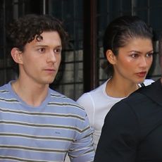 Zendaya wears a white T-shirt and linen pants and has slicked back short hair while holding hands with Tom Holland